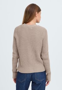 Beige knit sweater with textured pattern, ribbed cuffs, and hem. Short length and relaxed fit, styled with dark blue jeans.