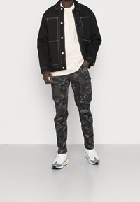 Black denim jacket with white stitching, cream t-shirt, camo cargo pants, and gray sneakers with neon accents. Pants feature pockets.