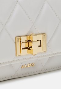 Přešívaná bílá kožená kabelka se zlatým geometrickým zavíráním a vyraženým logem "ALDO". Má šikmé švy a hladký povrch.