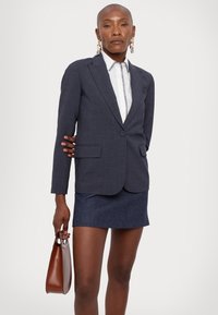 Dunkelgrauer maßgeschneiderter Blazer mit Revers und zwei Fronttaschen, getragen über einem weißen Hemd mit dekorativem Besatz und kombiniert mit einem blauen Rock.