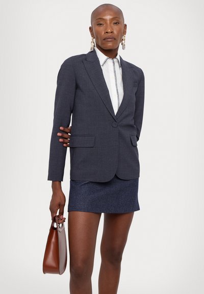Blazer su misura grigio scuro con rever a punta e due tasche frontali, indossato sopra una camicia bianca con rifiniture decorative e abbinato a una gonna blu.