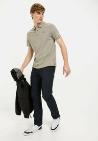 Lysegrå polo skjorte med krave og to knapper, parret med marineblå bukser og hvide sneakers, mens der holdes en sort jakke.