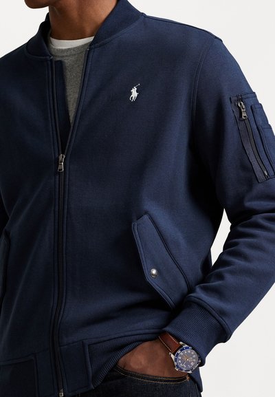 Jūras zilas krāsas džemperis ar rāvējslēdzēju un ribotu apkakli, sānu kabatām un izšūtu logotipu. Tam ir rāvējslēdzēja kabata uz piedurknes un gluda tekstūra.