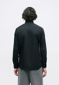 Cămașă lungă neagră cu mânecă lungă, guler clasic, textură netedă, mansete butonate și tiv curbat, purtată cu pantaloni gri.