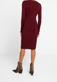 Robe en maille bordeaux à manches longues, silhouette ajustée et longueur mi-mollet. Associée à des bottines cavalières marron à motif de serpent.