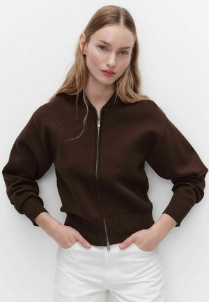 Jonge vrouw met lang blond haar, gekleed in een donkerbruine sweater met rits en witte broek, handen in de zakken, tegen een effen lichte achtergrond.