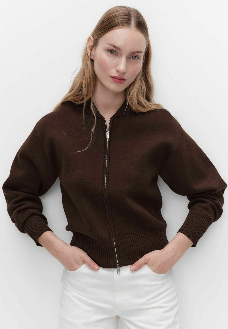 Jonge vrouw met lang blond haar, gekleed in een donkerbruine sweater met rits en witte broek, handen in de zakken, tegen een effen lichte achtergrond.