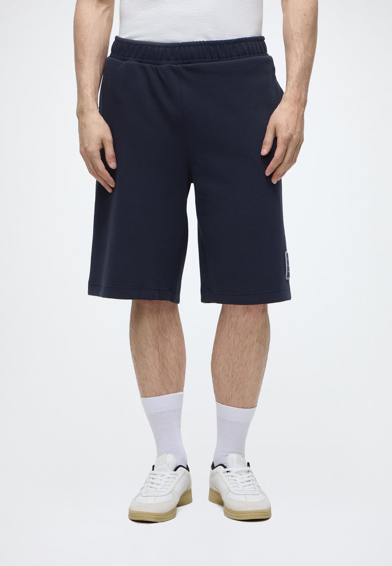 Homme portant un short bleu marine mi-genoux, des chaussettes blanches, des baskets blanches et un t-shirt blanc, debout les mains détendues le long du corps.