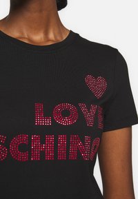 Svart t-shirt med kort ärm, som har stora röda rhinestone-text som stavar "LOVE" och "SCH" med en liten hjärt-accent, mjuk tygtextur.