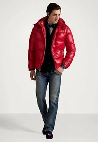 Vyras vilkintis blizgančią raudoną pūkinę striukę ant juodo megztinio ir marškinių su kaklaraiščiu, mėlynus džinsus ir tamsius loafers, žengiantis į priekį.