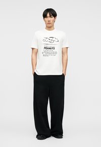 Jeune homme portant un T-shirt blanc avec un motif Peanuts mettant en scène Snoopy, associé à un pantalon noir ample et des chaussures noires, debout devant un fond uni.
