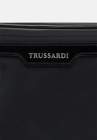 Svart Trussardi-plånbok med strukturerad tyg, med glansig svart detalj och präglad silverlogotyp på framsidan.