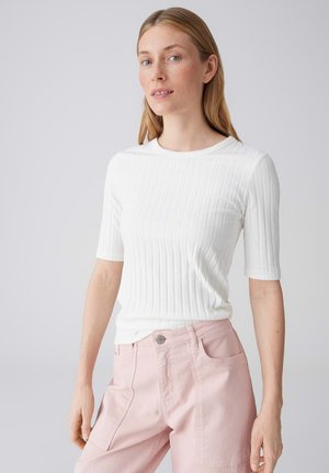 Donna dalla carnagione chiara e capelli biondi lisci indossa una maglietta bianca a maniche corte a costine e pantaloni rosa chiaro, in piedi davanti a uno sfondo semplice.