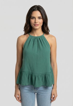 Femme aux longs cheveux foncés portant un haut vert sans manches à volants et un jean bleu clair, debout devant un fond gris clair uni.