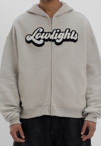 Světle šedá zipová mikina s hebkým černým logem "Lowlights". Má volný střih, žebrované manžety a kapuci na šňůrku.