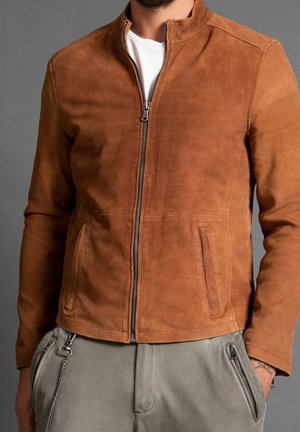 Hombre con una chaqueta marrón de ante con cremallera sobre una camisa blanca y pantalones gris claro con bolsillos con cremallera, de pie contra un fondo gris.