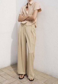 Pantaloni larghi in lino di un beige chiaro abbinati a una camicia a maniche corte con bottoni a righe nei colori pastello. Indossate sandali piatti neri.