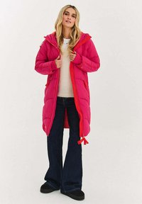 Nafouklý růžový kabát s kapucí, zapínáním na zip a stahovací šňůrkou. Má prošívanou texturu a kontrastní oranžové akcenty, doplněný tmavými džínami.
