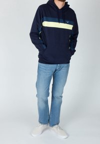 Felpa blu navy con un motivo a righe orizzontali in verde chiaro e blu scuro, dotata di tasca frontale e cappuccio con lacci, abbinata a jeans azzurri.