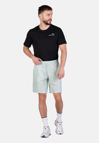 Pantaloni corti verde chiaro con una vita elastica e tasche laterali, abbinati a una maglietta nera a maniche corte e scarpe da ginnastica bianche.