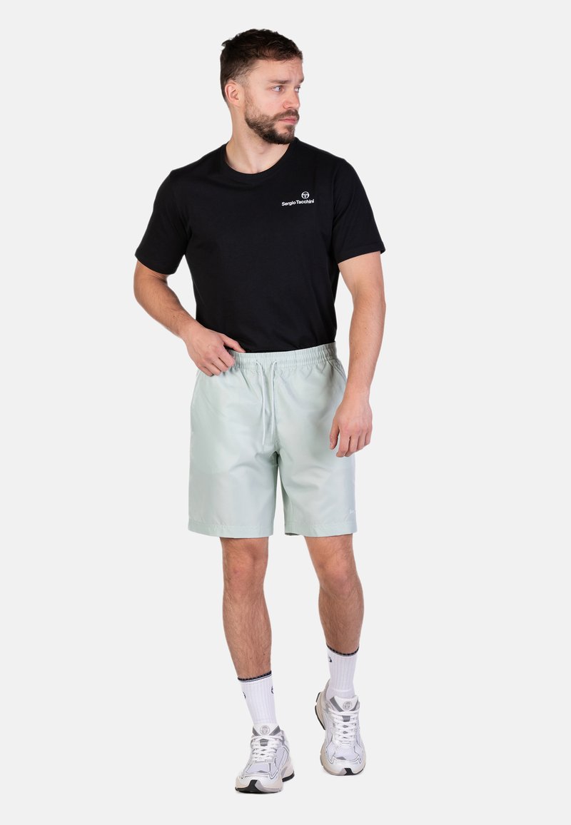 Pantaloni corti verde chiaro con una vita elastica e tasche laterali, abbinati a una maglietta nera a maniche corte e scarpe da ginnastica bianche.