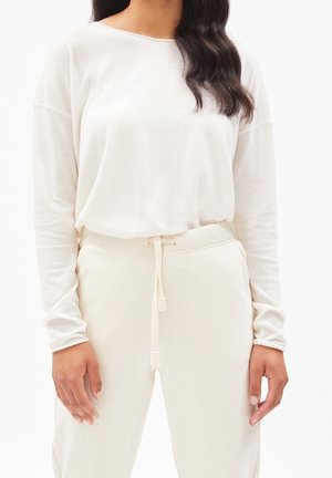 Femme portant un haut blanc à manches longues et un pantalon blanc assorti avec cordon de serrage, debout, les bras détendus le long du corps, devant un fond blanc.