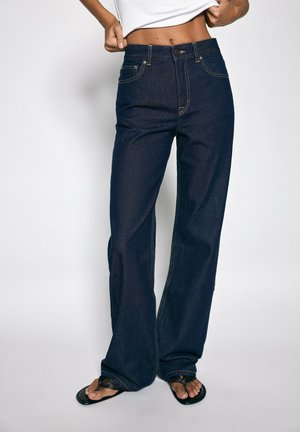 Personne portant un jean large bleu foncé et des sandales tongs noires, remontant un haut court blanc pour montrer le ventre.