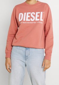Rosa Crewneck-Sweatshirt mit weißem "DIESEL"-Text und Slogan. Aus weichem Material gefertigt, mit gerippten Bündchen und Saum, lockere Passform. Hellblaue Jeans.