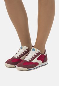 Rote und weiße Wildledersneaker mit niedriger Profilhöhe, ausgestattet mit weißen Schnürsenkeln, einem blauen Logo auf der Zunge und einer strukturierten Gummisohle.