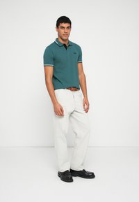 Türkizkék galléros póló fekete gallérral és csíkos mandzsettákkal, világos színű nadrággal és fekete cipővel párosítva. A modell egyenesen áll, karjai lazán maga mellett.