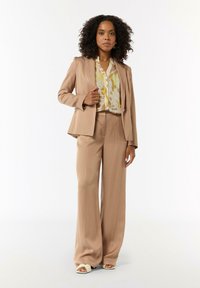 Beige tailored suit with long, loose pants and fitted jacket. Underneath, a light blouse featuring a yellow and green abstract pattern.