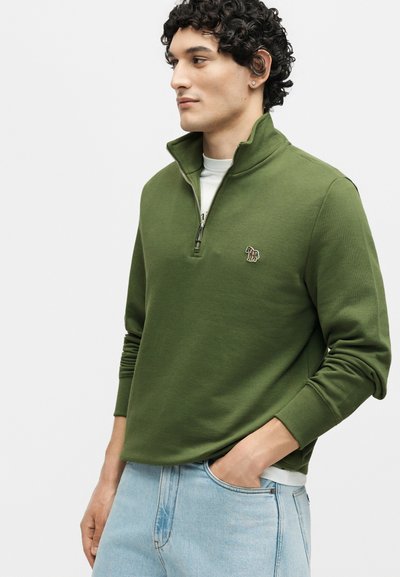 Άντρας με σγουρά μαλλιά, που φοράει ένα χακί φούτερ με φερμουάρ στο τέταρτο και μικρό κεντητό λογότυπο ζέβρας και ανοιχτό μπλε τζιν.