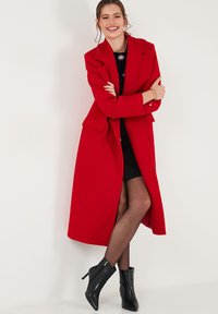 Red wool overcoat with notched lapels and side pockets, worn over a black dress. Black ankle boots complete the outfit.
