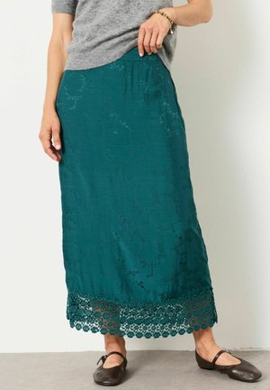 Kvinde iført en teal maxi nederdel med blomsterbroderi og blonderkant kombineret med en grå sweater og mørkebrune flade sko.