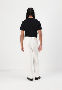 Černé tričko s krátkým rukávem doplněné bílými kalhotami s rovnými nohavicemi, ukazující minimalistický design a lehkou, jemnou texturu. Černé boty dotvářejí celý outfit.