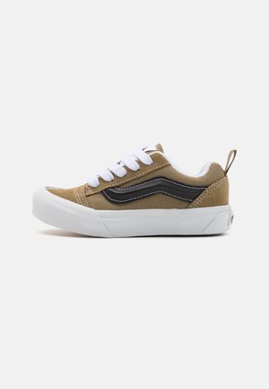 Baskets en toile beige avec une semelle en caoutchouc blanc, une bande latérale noire et des lacets blancs. Comprend une languette à l'arrière et des détails de couture.