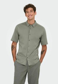 Chemise à manches courtes en tissu vert clair, avec un col pointu et des boutons contrastants. Texture lisse, coupe ajustée.