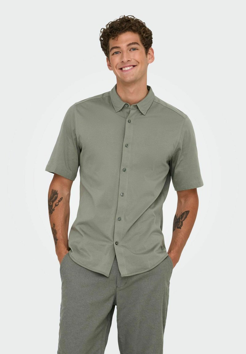 Chemise à manches courtes en tissu vert clair, avec un col pointu et des boutons contrastants. Texture lisse, coupe ajustée.