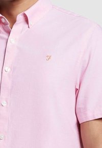 Camicia button-up rosa a maniche corte realizzata in tessuto leggero, con un discreto logo dorato sul petto e chiusura con bottoni standard.