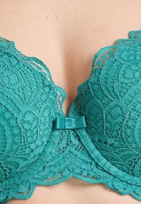 Sostenitore in pizzo teal con motivi floreali intricati, bordo scallopato e dettaglio in satin a forma di fiocco al centro davanti. Design delicato e testurizzato.