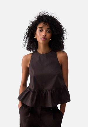 Donna con capelli ricci che indossa orecchini dorati e una maglia nera senza maniche con un orlo arricciato, in piedi con le mani in tasca.