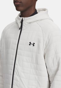Veste matelassée blanche avec capuche, fermeture éclair intégrale et accent noir à l'avant. Fabriquée en matériau synthétique lisse avec un motif texturé.