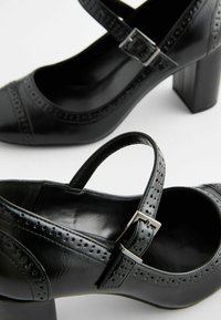 Zapatos de tacón alto de cuero negro con punta redondeada, correa en el tobillo y detalles de brogue. Tacón cuadrado. Textura suave con acabado brillante.