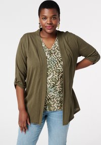 Cardigan vert olive à manches trois-quarts porté sur un haut à motifs vert, beige et bleu. Un jean bleu clair complète la tenue.