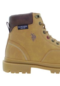 Stivale da trekking beige con lacci, colletto imbottito marrone, suola spessa in gomma, etichetta U.S. Polo Assn. e logo del giocatore di polo sul lato.