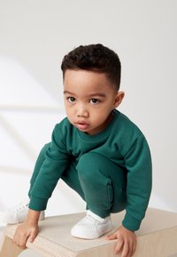 Grüner Sweatshirt und passende Jogginghose aus weichem Stoff. Das Kind trägt weiße Turnschuhe und hockt auf einer Holzoberfläche.