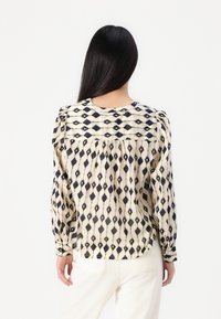 Long-sleeve blouse in cream with black diamond pattern, featuring gathered detailing on shoulders and back, and buttoned cuffs.