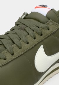 Zaļas sporta kedas ar teksturētu olīvzaļo augšdaļu, baltu Swoosh logo un atbilstošiem auklām. Iespējo mīkstināta mēle ar kontrastējošu sarkanu logotipu.