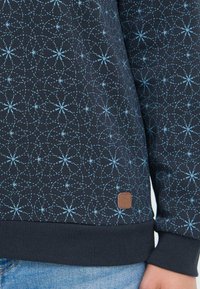 Sweatshirt bleu marine avec un design géométrique en bleu clair, des poignets et un ourlet côtelés, et un petit patch logo marron sur le côté gauche.