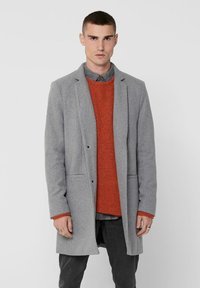 Grau Wollmantel mit Revers, zwei vorderen Taschen und Knopfverschluss, kombiniert mit einem orangefarbenen strukturierten Pullover und dunklen Hosen.